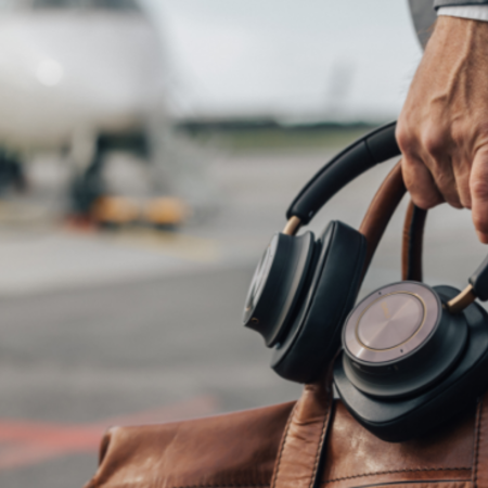 Eine Hand hält eine Ledertasche und DALI-Kopfhörer, ein Flugzeug ist im Hintergrund erkennbar