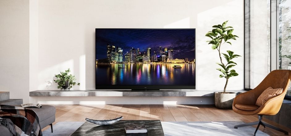 Produktneuheit Panasonic OLED Serie | OLED TV-Modell in einem modern eingerichtetem Wohnzimmer mit einer grauen Couch, einem gelben Sessel und zwei Zimmerpflanzen, hell-erleuchtet