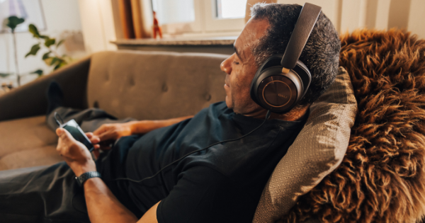 Dali IO-12 Kopfhörer Neuheit | Ein Mann liegt auf einer grauen Couch mit aufgesetzten DALI-Kopfhörern und einem MP3-Player in der Hand, Ansicht im Seitenprofil mit warmen Licht-Einfall